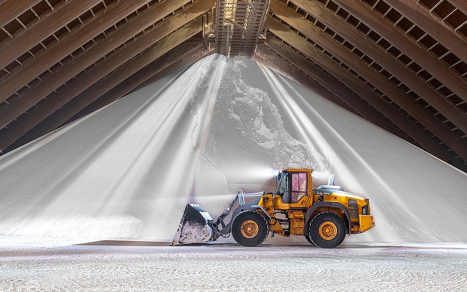 Ein gelber Bagger schaufelt in einer großen Lagerhalle einen riesigen Haufen Salz. Lichtstrahlen fallen durch die Dachstruktur und beleuchten das weiße Salz, was eine dramatische Szenerie schafft.