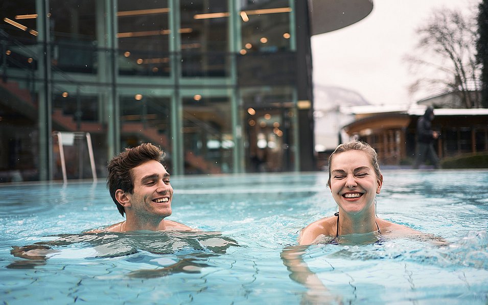  Ein Mann und eine Frau genießen lachend ein Bad in einem Außenbecken einer modernen Therme. Im Hintergrund sind Glasfassaden und warme Beleuchtung zu sehen, während es draußen kühl wirkt, möglicherweise Winter.