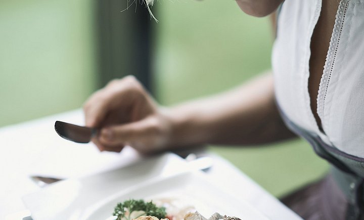 Eine Frau in Tracht, von schräg oben gesehen, hält Gabel und Messer gerade in der Hand, um die vor ihr auf dem Tisch stehende Speise zu genießen.