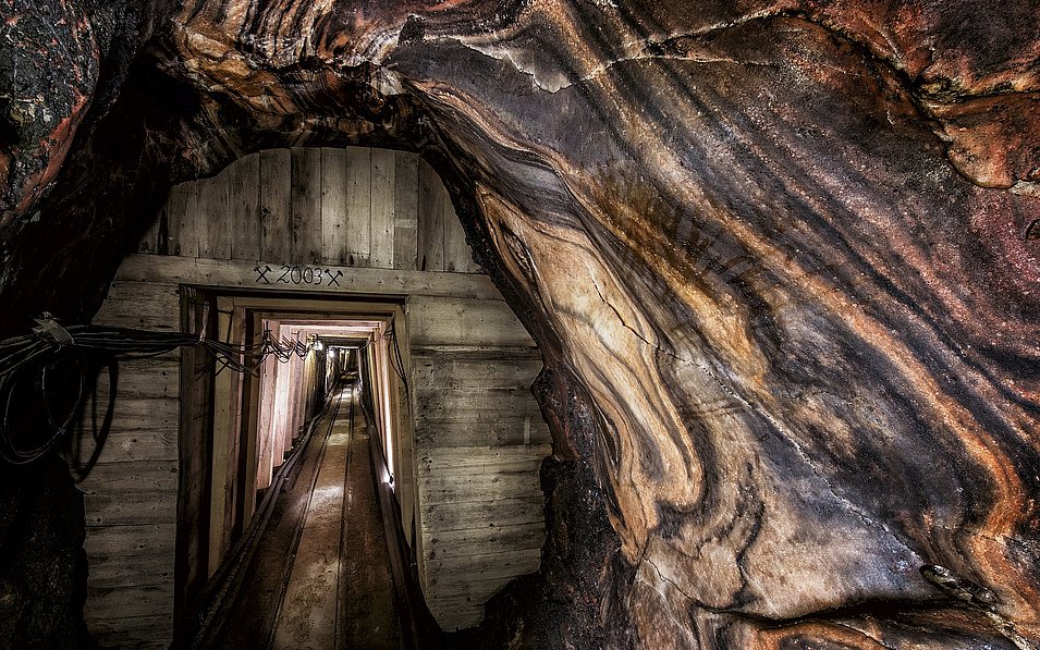 Ein schmaler Tunnel, dessen Eingang aus Holzbalken besteht, führt tief in das Innere eines Salzbergwerks. Die Wände des Tunnels sind von dunklen, wellenförmigen Salzgesteinsschichten geprägt, die in warmen Farben schimmern.