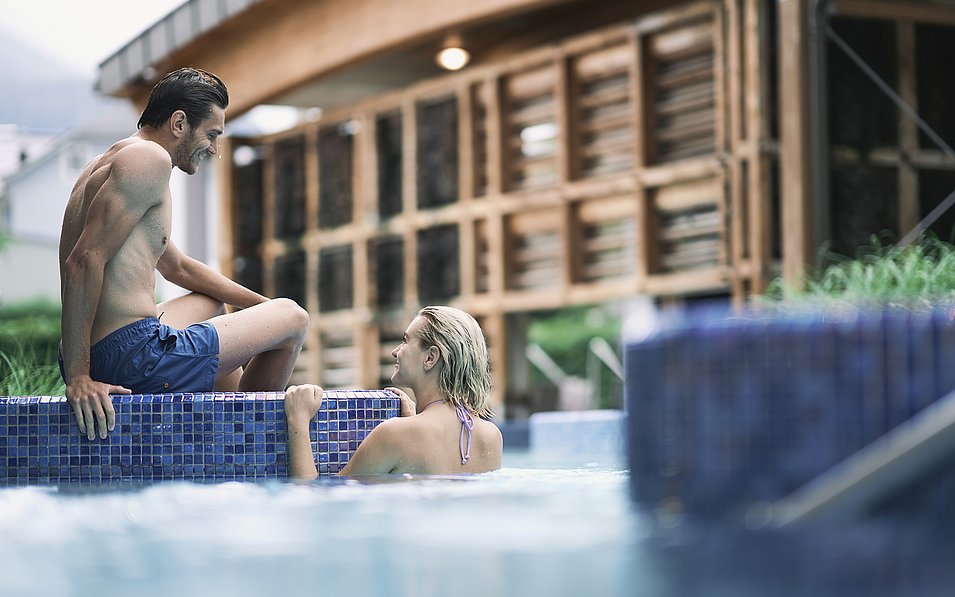 Ein Mann sitzt am Beckenrand, während eine Frau im Wasser steht und zu ihm aufblickt. Im Hintergrund ist ein Holzgebäude einer Therme zu sehen.