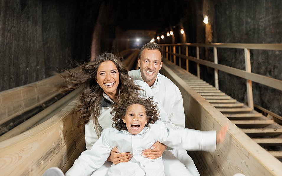 Eine fröhliche Familie in weißen Schutzanzügen rutscht im Bergwerk gemeinsam eine Holzrutsche hinunter. Sie werden von warmen Lichtern entlang des Tunnels beleuchtet. Die drei Personen strahlen pure Freude aus.