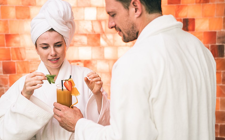 A man and a woman in bathrobes inside the SkyLounge wellness area on the roof of the Hotel Royal in the EurothermenResort Bad Ischl in the Salzkammergut, the man holding a drink from which the woman is removing mint leaves.